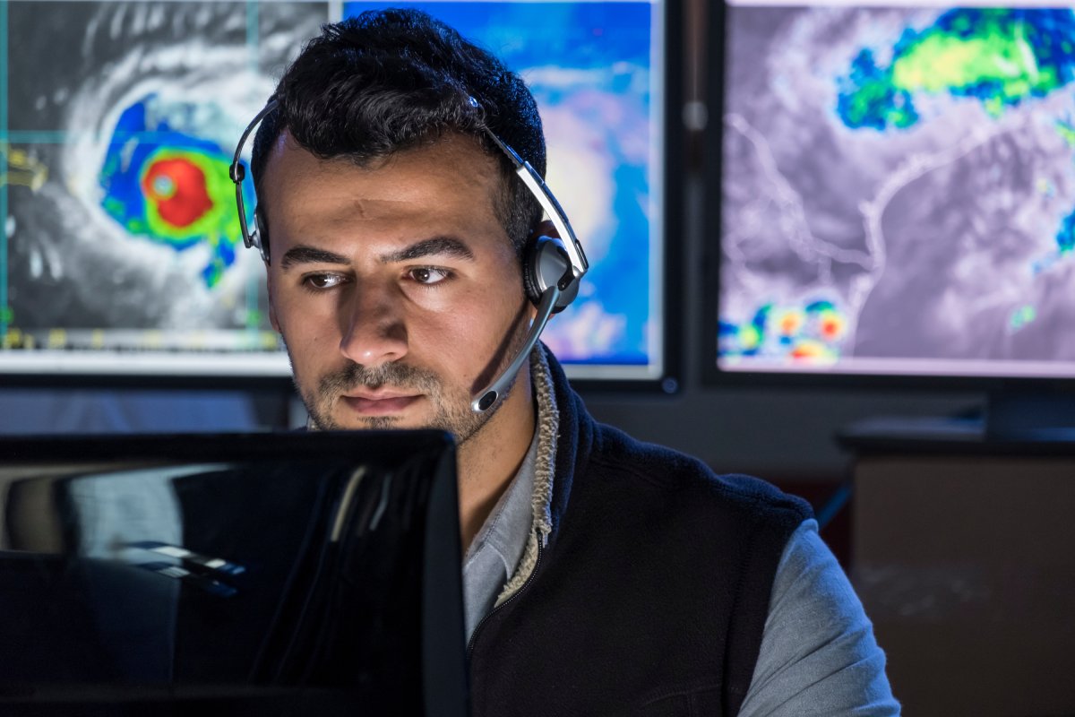 A man with a headset looking concerned with screens showing storms behind him.